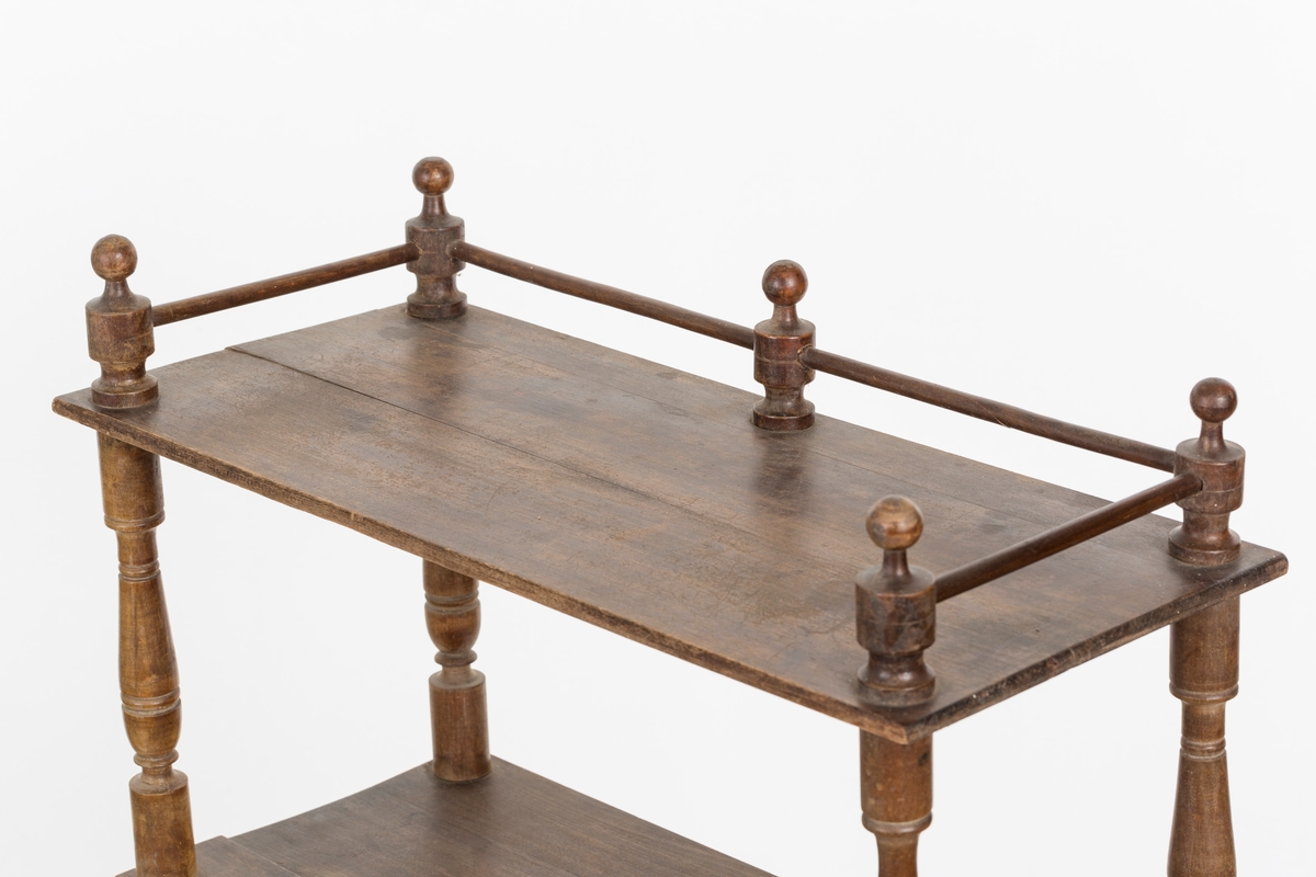 Hylle med 4 hyller. Dreide stolper mellom hyllene. Mangler ett bein. Sprosser øverst. Lakkert i mørk brunt.