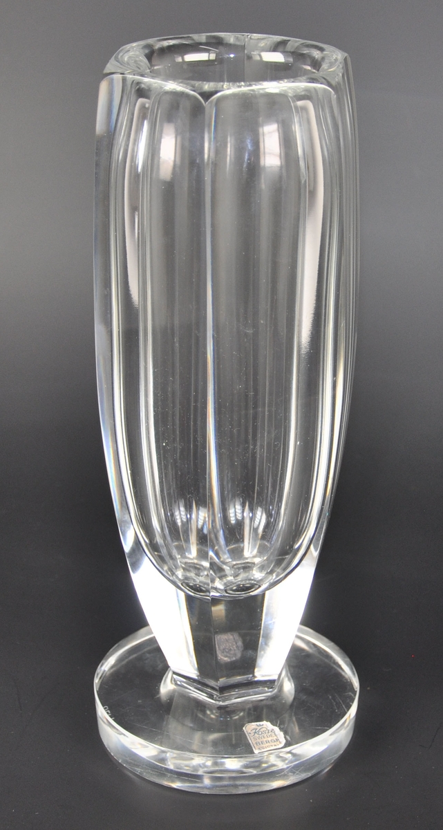 Kantet blomstervase av glass med rund fot. Klistrelapp med produsentmerke på vasen.