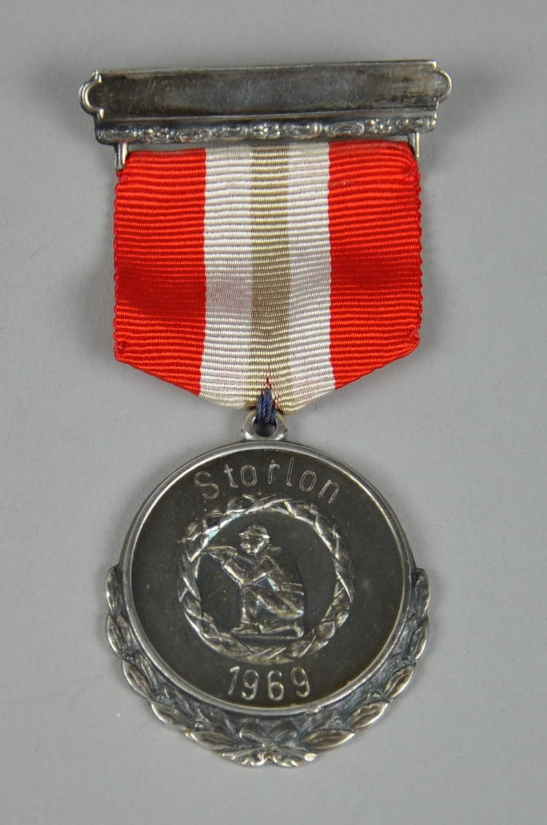 Medalje med laurbærkrans rundt medaljongen og skiskytter som sitter på kne og sikter med geværet. Krans rundt skytteren. Nål med bånd i rødt, hvit og blått (blå farge er falmet).