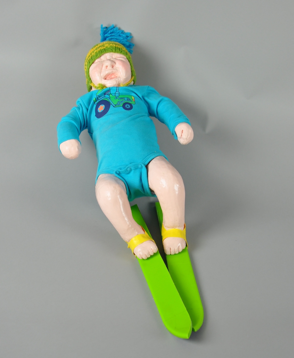 Dukke med ski på beina. Dukken har på seg en grønn og gul strikket topplue med blå dusk og en blå body med "Moods of Norway"-traktor. Skiene er grønne med gule bindinger av tape.  Dukken kan være inspirert av statuen "Sinnataggen" i Vigelandsparken.