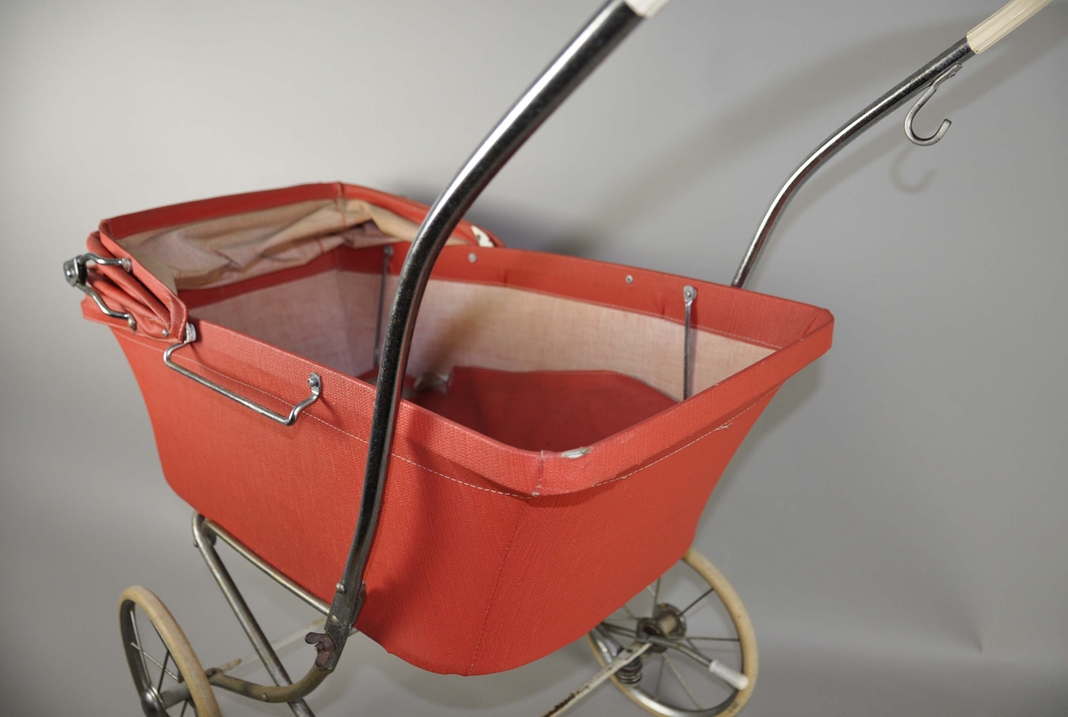 Dukkevogn med hjemmelaget madrass, dyne og pute. Vognen er av rød polyester med sammenleggbar kalesje. På sidene er det håndtak til å feste et løst vogndekke. Håndtak av kromt metall går ned på sidene av vognen, øverste del er lagt med hvit plast. Hjulene er hvite med eiker, ved høyre hjul er det en bremsespenne. Understellet på vognen er en lys treplate. På høyre håndtak er det satt på en refleks, av nyere tid.