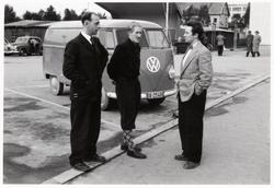Tre menn på fortau. VW-buss i bakgrunnen.