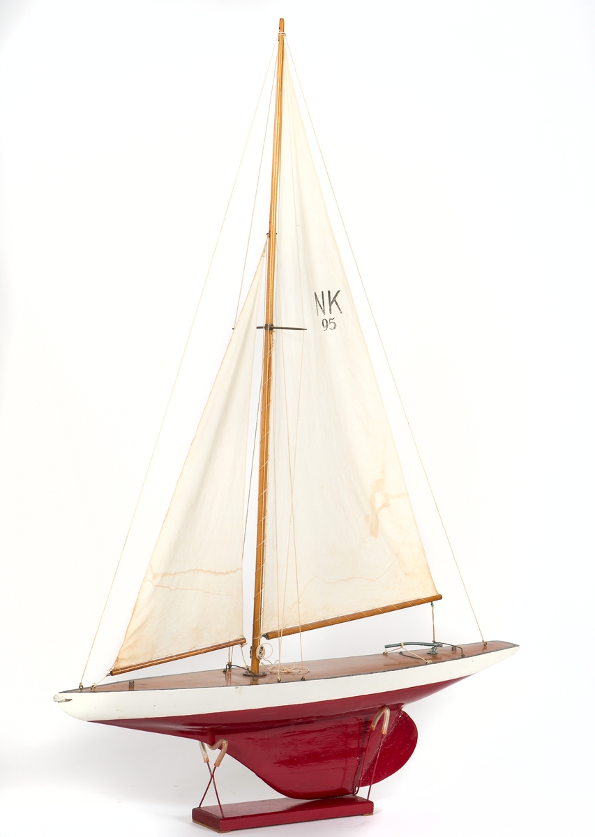 Modell av segelbåt, leksak. Tillverkad av NK senast 1935. 
Vitt skrov med röd bottenmålning. Fernissat nåtat däck och blyköl.