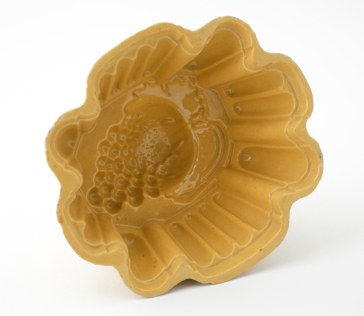 Aladåbform i keramik. 
Oval veckad form på fot, med inpressad dekor i form av druvklase i botten. Honungsgul glasyr.