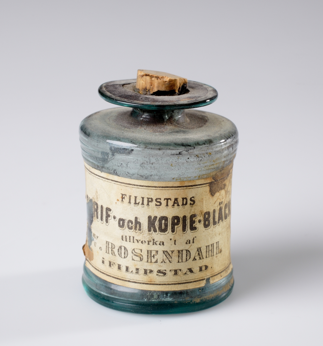 Bläckflaska av blåtonat glas. 
Cylindrisk modell med troligen sekundär propp av kork. Etikett runt flaskan med text: "Filipstads Skrif- och Kopie-bläck, tillverkadt af W. Rosendahl i Filipstad".