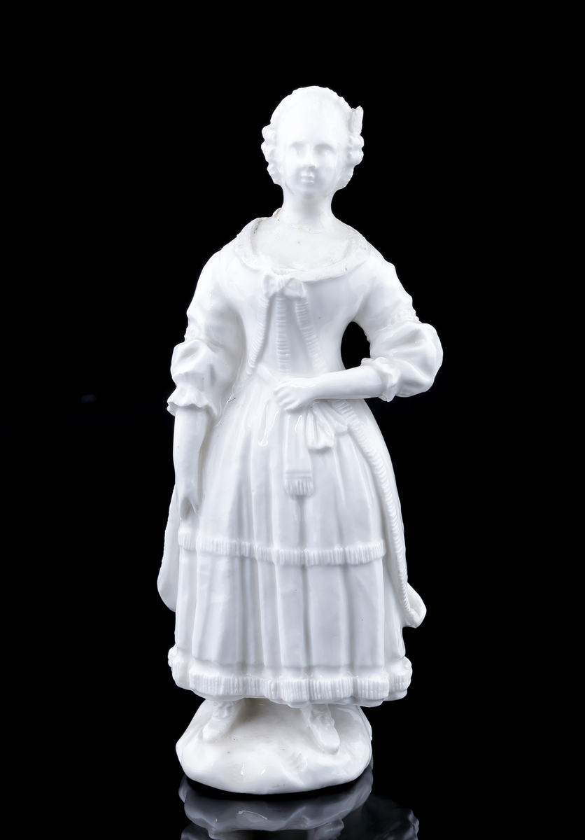 Figurin i vitt porslin. 
Kvinna klädd i 1700-talskläder med kappa. Mycket otydlig signering i botten. 
Troligen Marieberg, möjligen Rörstrand.