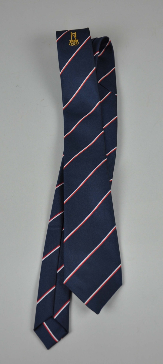 Blått slips  med NOK/NIFsin  logo og  de olympiske ringene innvevd  i lys  sennepsgult,plassert midt på slipset.Diagonale striper i hvitt og rødt.To like .