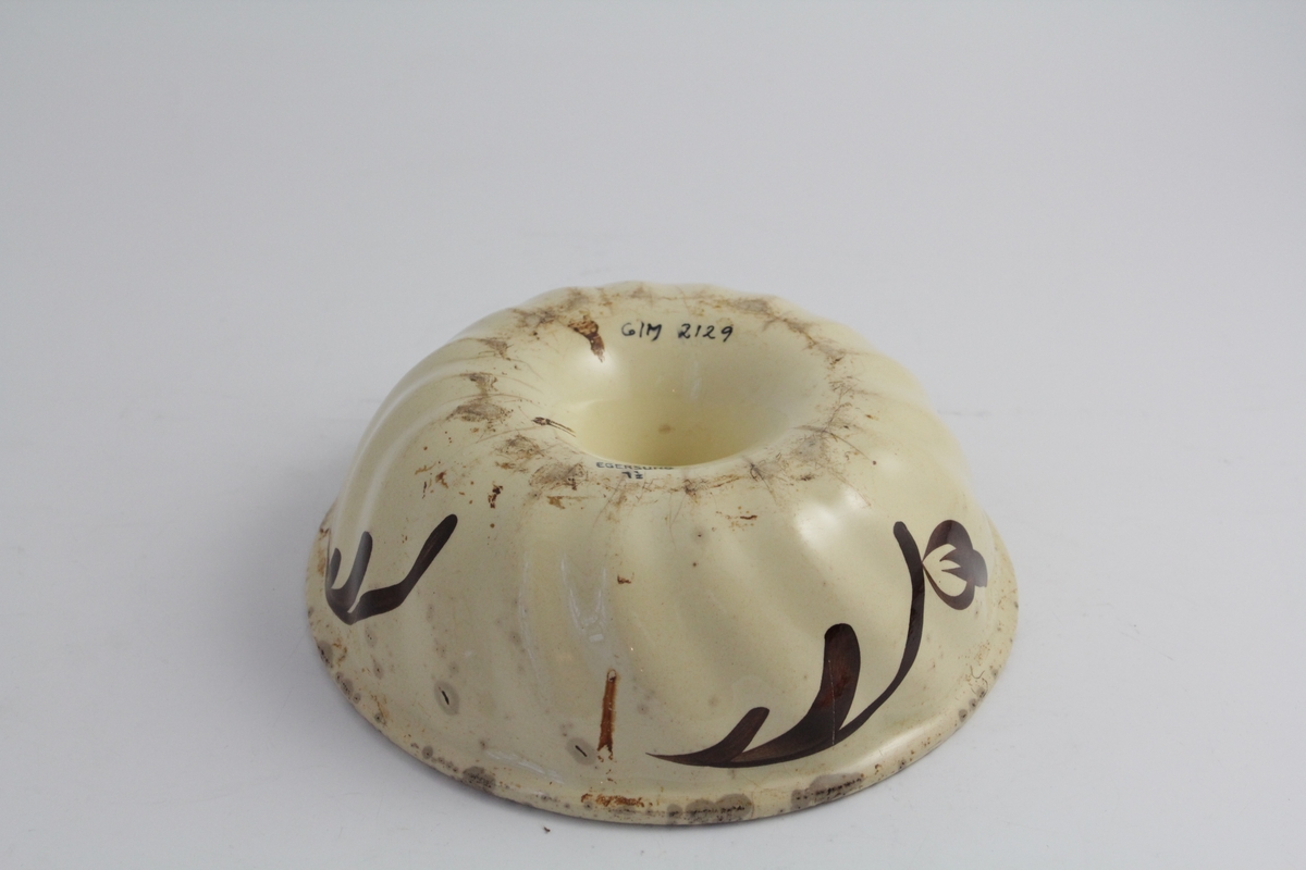 Rund form i ildfast steintøy. Kremgul farge, med brun dekor. Brune stiliserte blomsterkvister på sidene. Rommer halvannen liter.