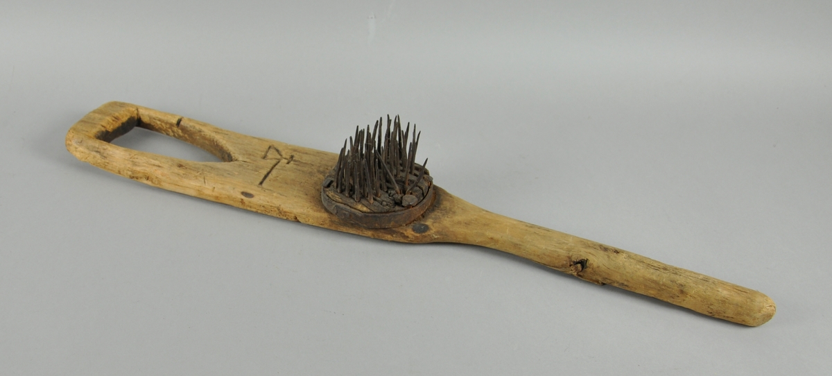 Linhekle som har et håndtak i den ene enden, den andre enden er smal og spiss. På midten av heklen er det en rundt kloss med stålspiker. Det er skåret ut et merke på brettet bestående av en trekant, en strek og et kors.