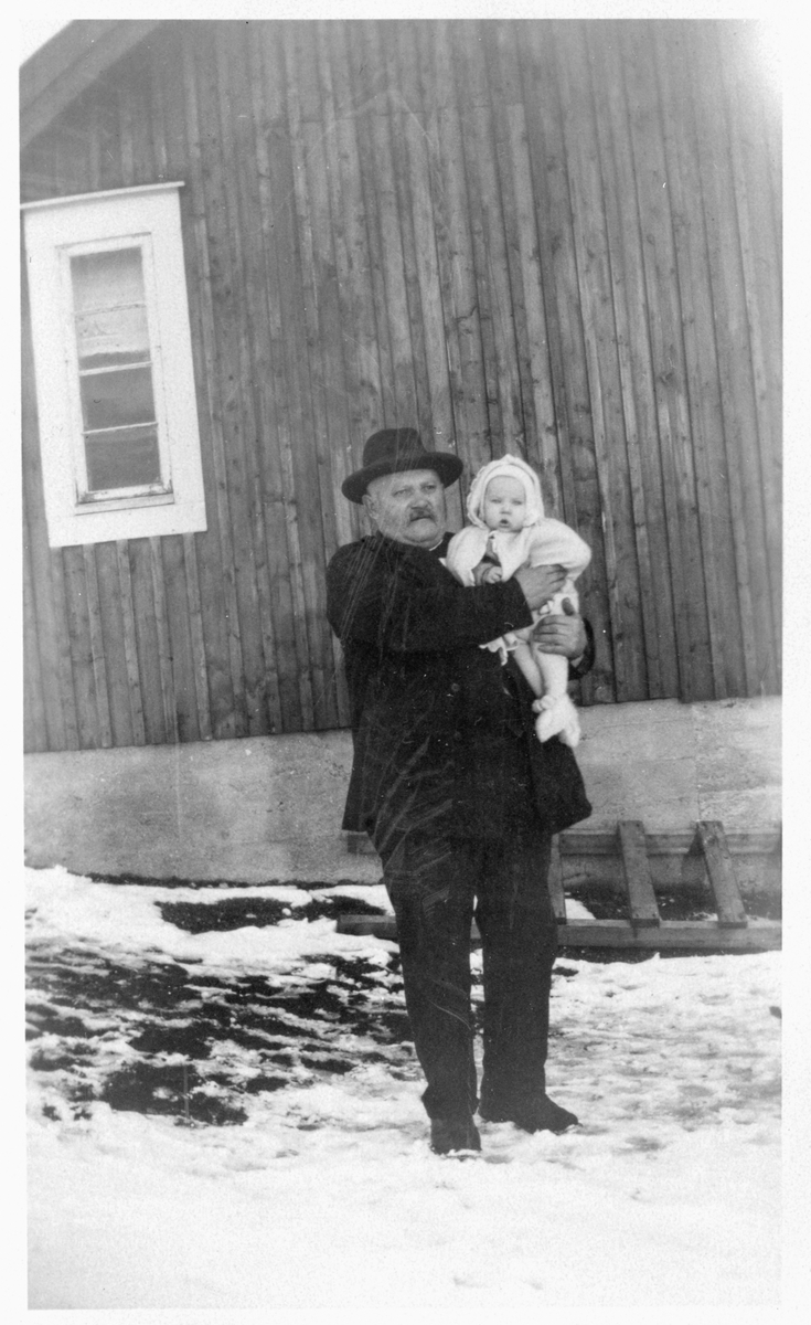 Mann med barn på armen, fotografert utendørs foran en husvegg.