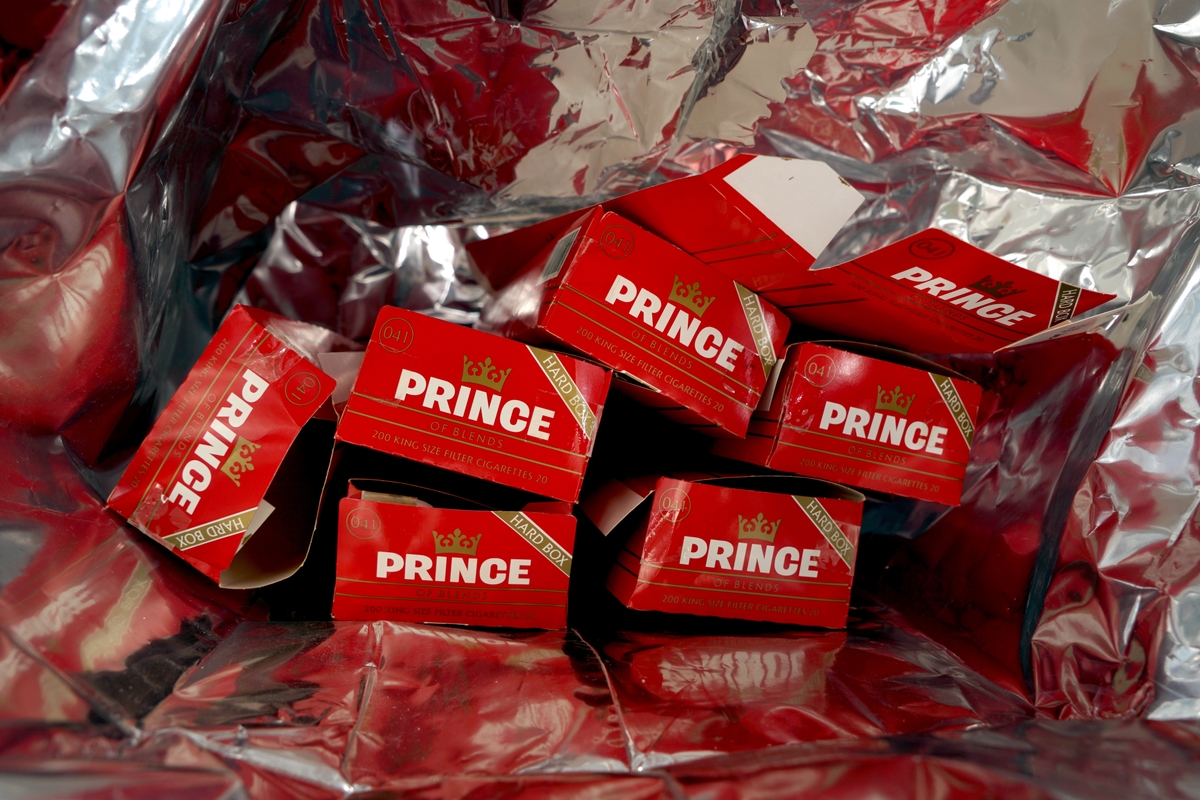 En sølvfarget plastsekk inneholdende sju brettede sigarettkartonger av merket Prince.
Ved siden av , 148 ubrettede sigarettkartonger i en bærepose fra Tiedemanns.