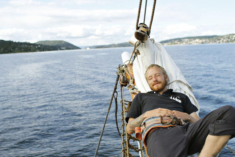 Mann slapper av ombord skonnerten "Svanen", blått vann, land og klar himmel i bakgrunnen. (Foto/Photo)