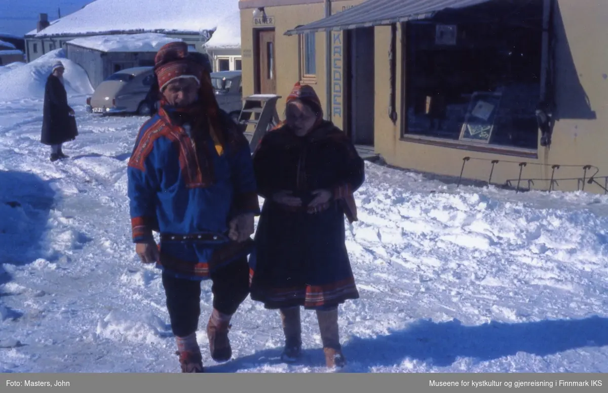 Honningsvåg. En samisk dame og en samisk mann i kofte. I bakgrunnen skredderbutikken. 1962/63.