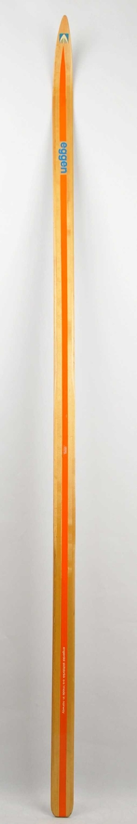 Langrennski i tre, lakkera trekvit overflate, oransje stripe på overside og på sidekantar. Emblem på skitupp. 