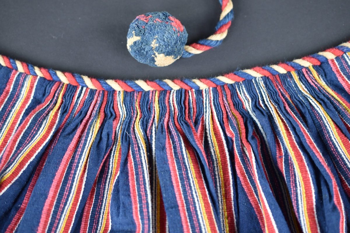 Förkläde av bomull, randat i rött, gult och vitt mot mörkblå botten. Virkad uddspets i blått, rött och vitt längs nederkanten. Förklädet rynkat mot en linning bestående av en röd-vit-blå snodd som även är midjeband med bollar i ändarna.