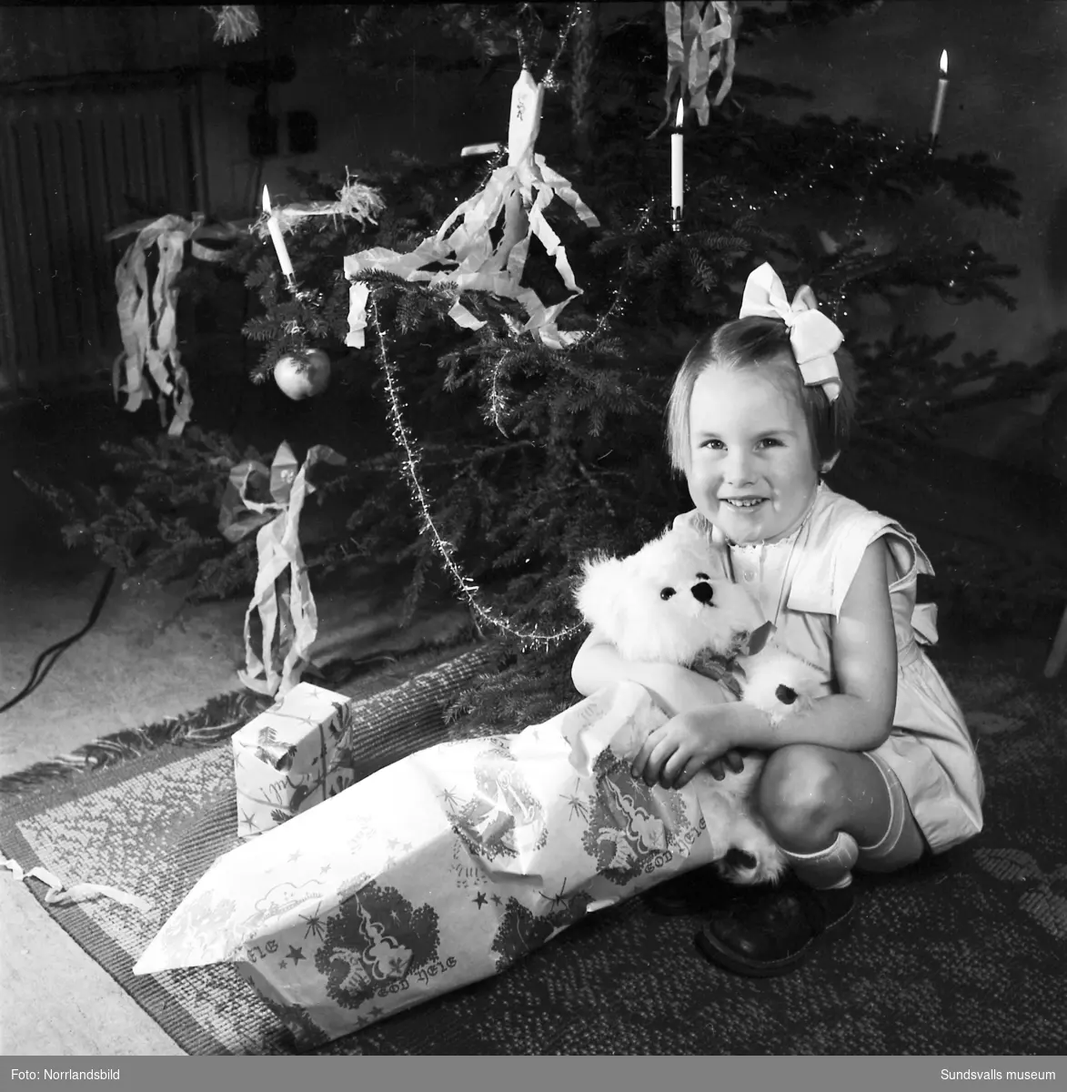 En liten flicka, Lena Tranberg, sitter framför julgranen med en nyöppnad present - en leksakshund.
