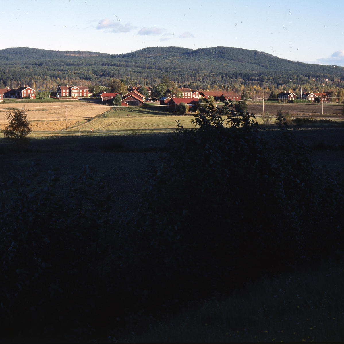 Norrvåga by i Järvsö, med skugga o påfallande sol, 6 september 2001. De röda, stora hälsingegårdarna ligger som på rad med bergen i bakgrunden. Framför breder ett jordbrukslandskap ut sig.