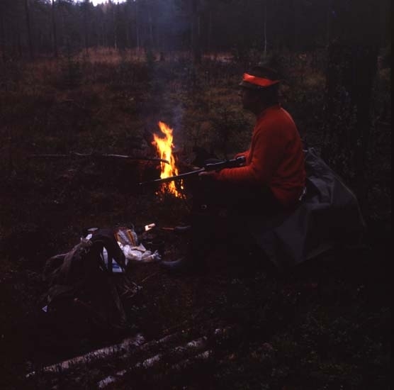 Älgjakt i Arbråskogarna 13 oktober 1980. En jägare med gevär sitter vid en eld på en myr. Bredvid sig har han en ryggsäck.
