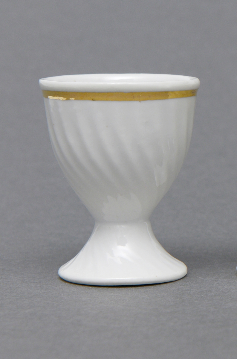 Eggeglass av porselen med hvit glasur. Rund stett og konisk formet klokke. Gullstripe rundt kanten.