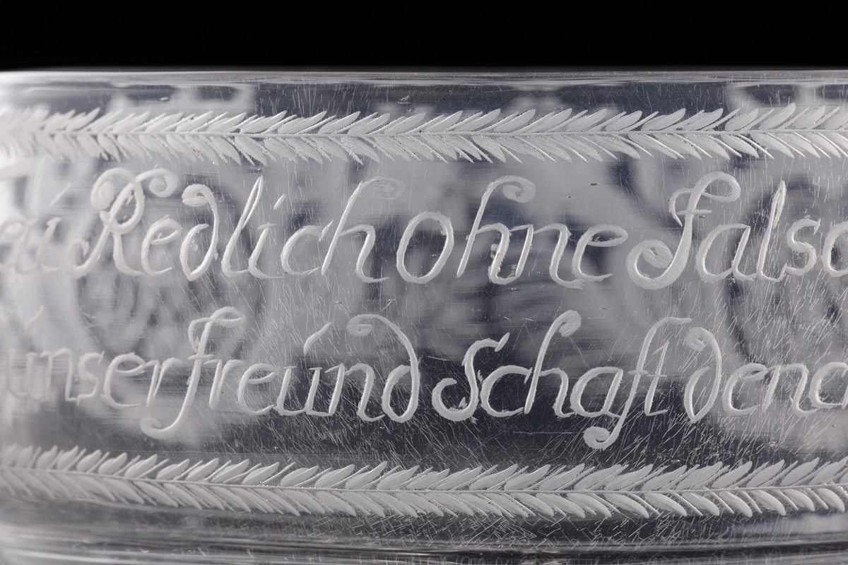 Skål av glas med hänklar. 
Graverad dekor med två spegelmonogram under friherrekronor och tysk text: "Treu Redlich ohne Falsch und Schein, Soll unser FreundSchaft dench Spruch Sein". 
Vilar på tre kulfötter.