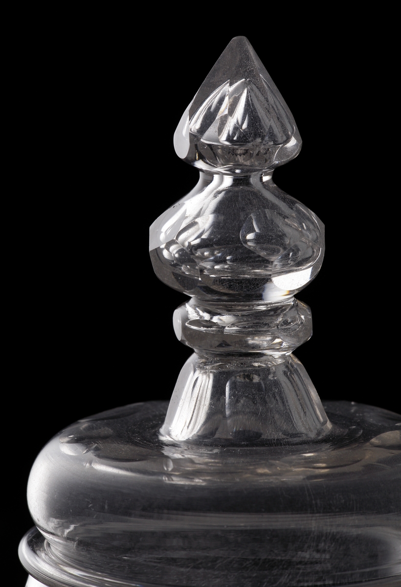 Pokal av glas, med lock. 
Slipat och graverat mönster med kungligt spegelmonogram: "FR"* i kartusch under krona. Invikt kant på foten.

*"FR" (Fredericus Rex= Fredrik I av Sverige, reg. 1720-1751).