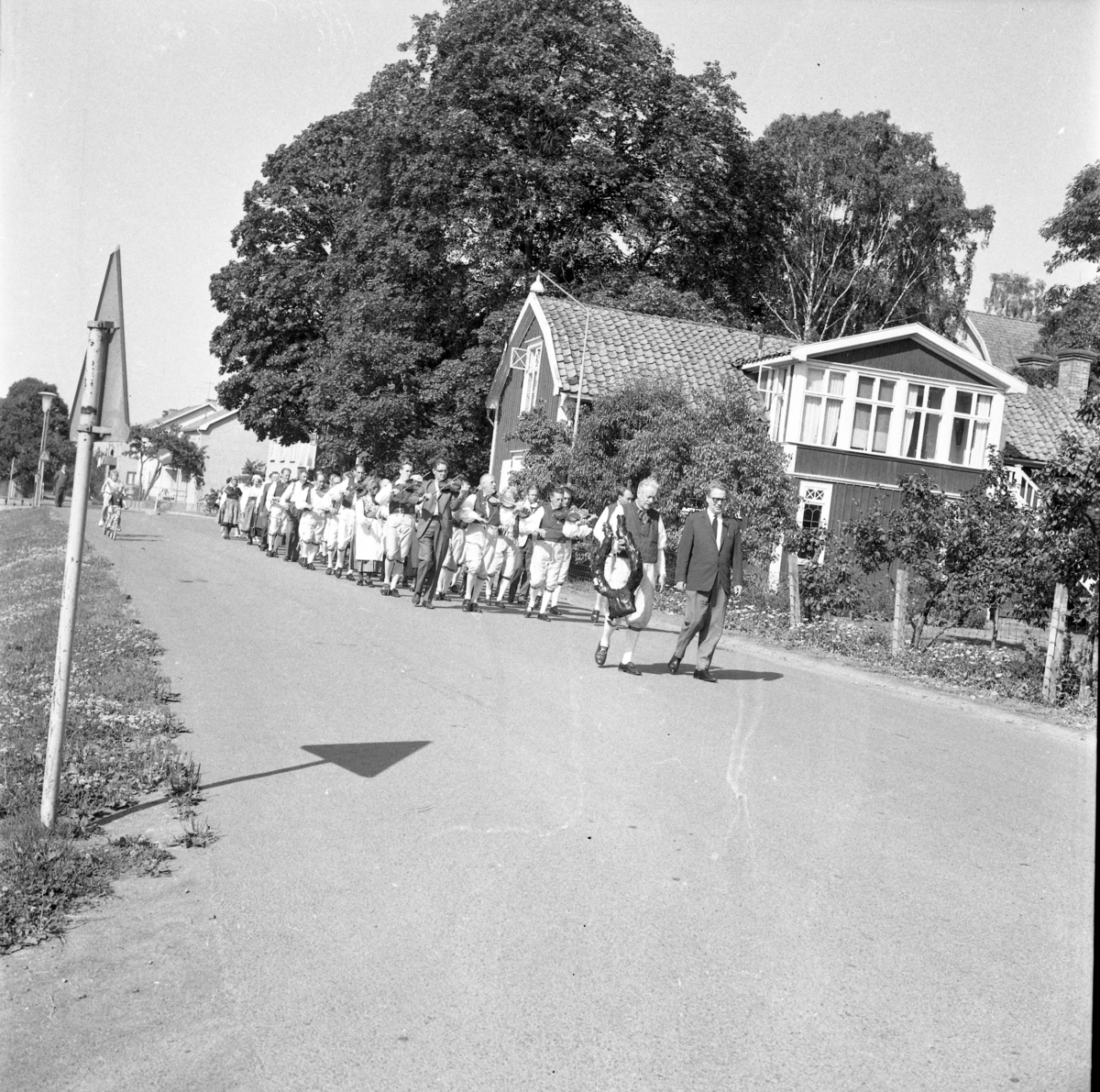 Södermanlands spelmanslag marscherar Gärdesvägen söderut, passerar Ribbagården, mot Gränna kyrka för att lägga en krans på "Blinde Jannes" grav, juli 1964. I täten går riksspeleman Gustaf Wetter med Tage Grennfelt till höger.