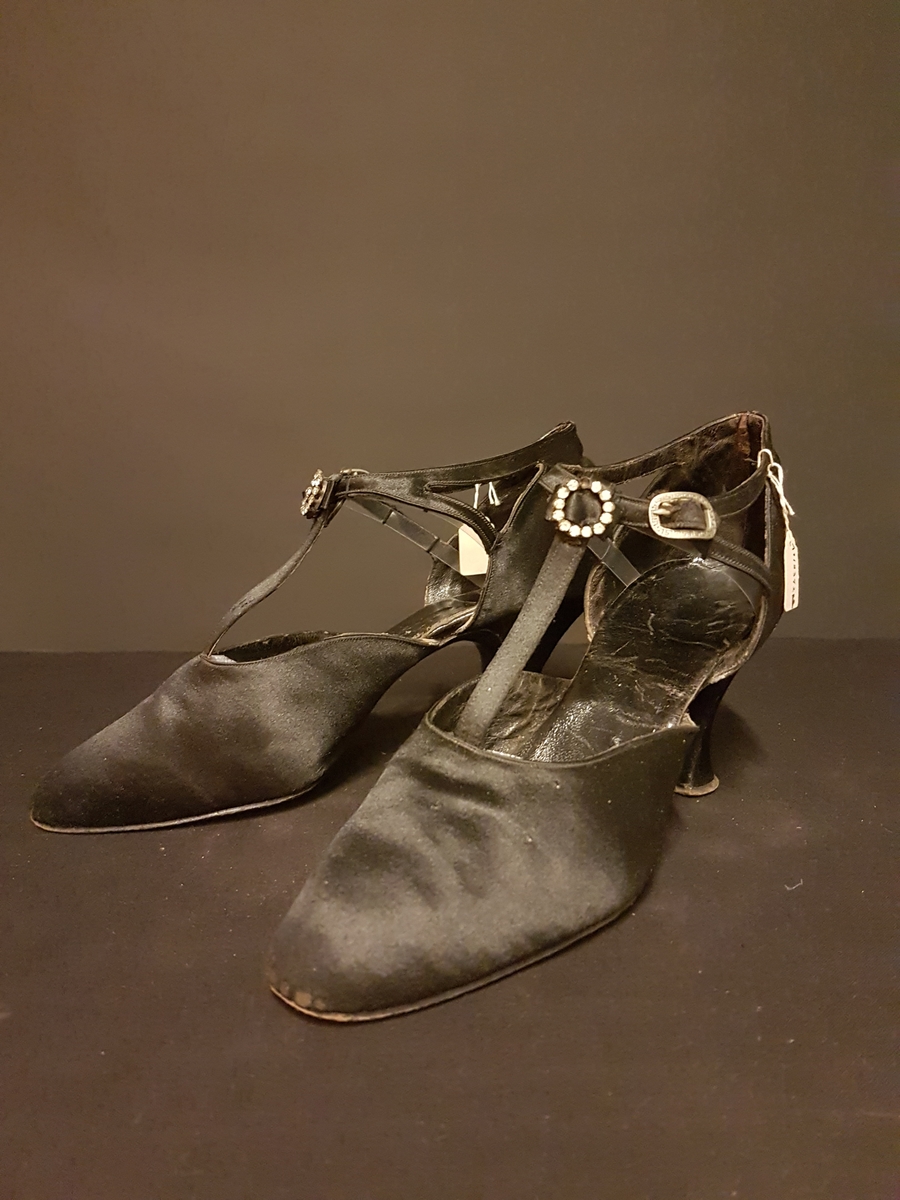 Ett par aftonskor i svart siden, klack, t-sleif spänne med meanderbård samt ett skospänne bestående av en ring med strasstenar.