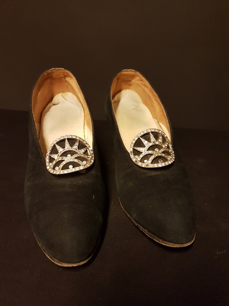 Pumps i svart mocka med klack och skospänne med strasstenar. I bägge skorna en mjuk extra sula.