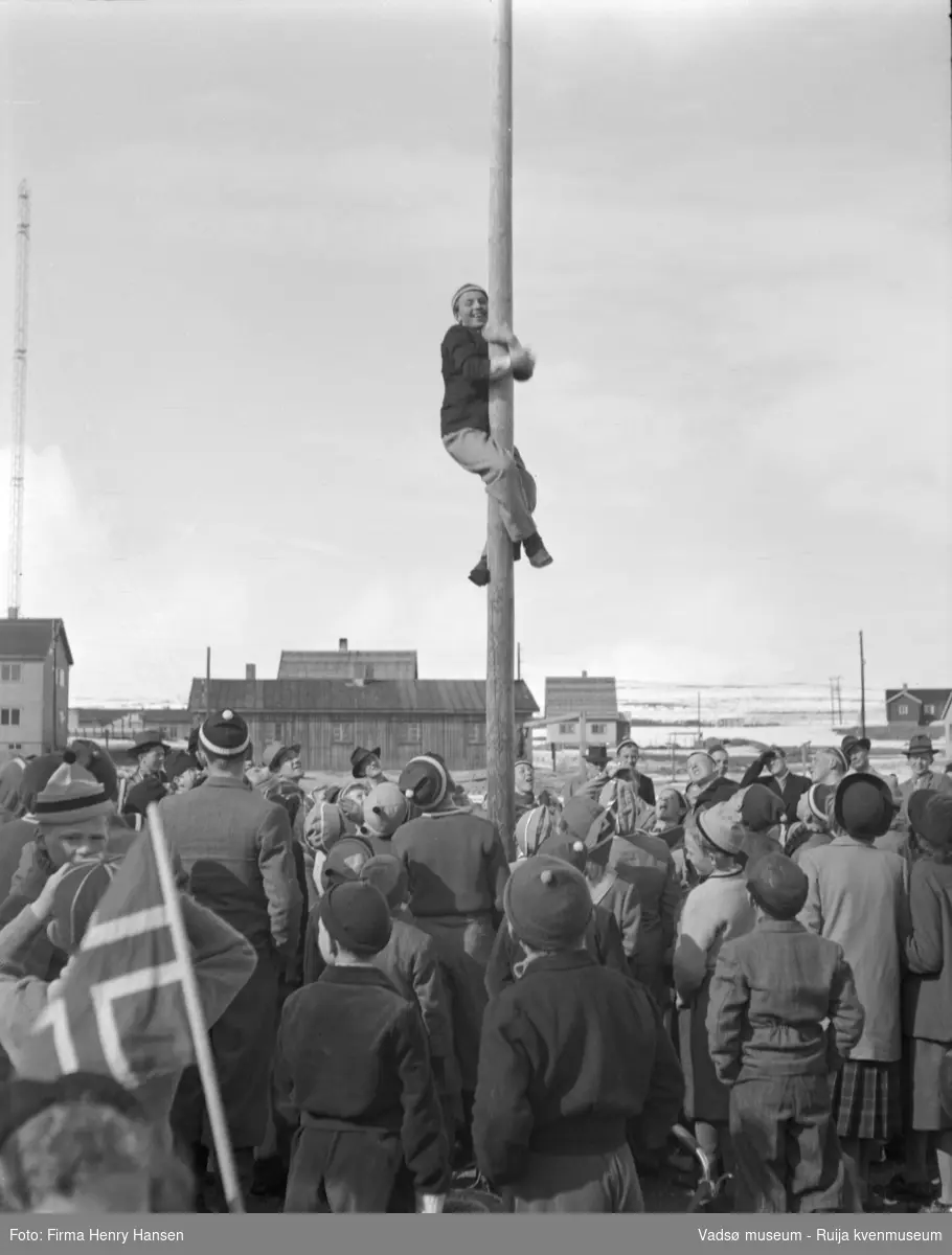 Vadsø 17 mai 1952. folk er samlet til lek og konkurranser der idrettshallen Vadsøhallen ligger idag (Brodtkorpsgate til venstre, hus langs Idrettsveien ovenfor). Ung gutt i topplue høyt i en flaggstang. Klatrekonkurranse med tilskuere. En av Finnmark kringkasters radomaster til venstre i bildet.