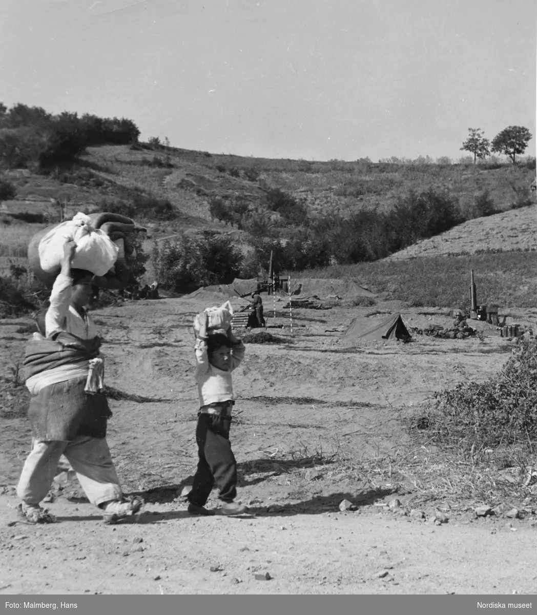 Koreakriget. I förgrunden koreanska flyktingar (ett barn och en kvinna) så bär sin packning på huvudet. I bakgrunden en artilleriposition med FN-trupper, kanoner och tält.