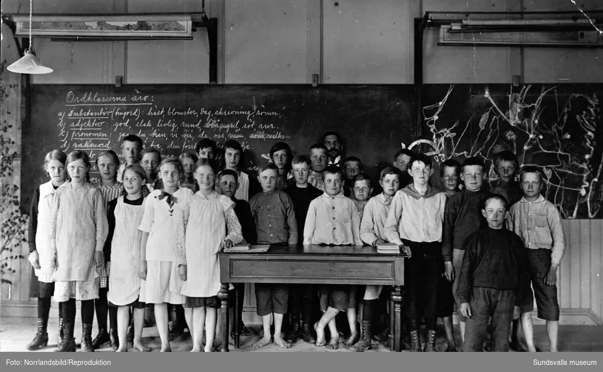 Klassfoto, möjligen skolavslutning. Barnen står uppradade framför svarta tavlan, en del har kängor andra är barfota. Reprofotografi för S. Nordenström, HSB.