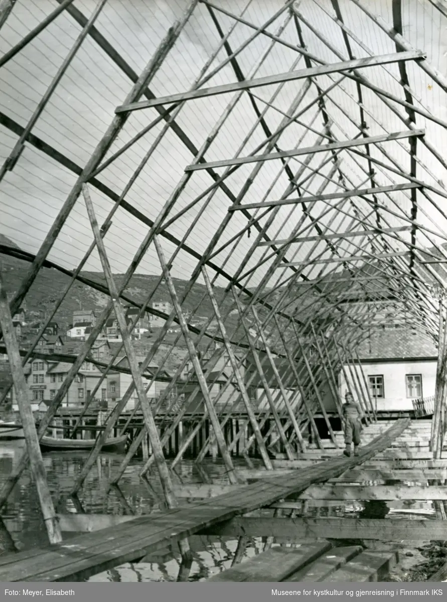 Honningsvåg. En gutt går på planker inne i en stor fiskehjell. Bebyggelsen ved Elvedalen ses i bakgrunn. 1940.