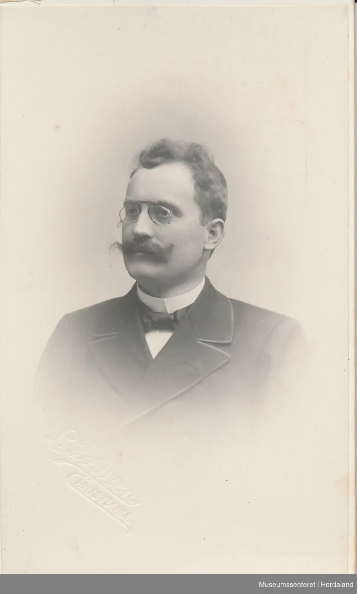 portrettfotografi av mann med bart, briller, dress, kvit stivesnipp og sløyfe