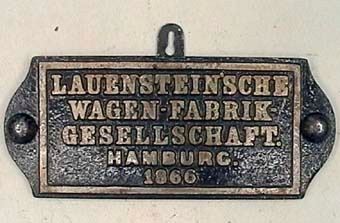 Rektangulär liggande skylt av svartmålat järn med rundade kortsidor. Silverfärgad text: "LAUENSTEIN'SCHE WAGEN-FABRIK-GESELLSCHAFT. HAMBURG. 1866".