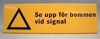 Rektangulär plåtskylt med svart text på gul botten:
"Se upp för bommen vid signal"
Till vänster om den svarta texten finns även en svart ofylld varningstriangel med basen nedåt.