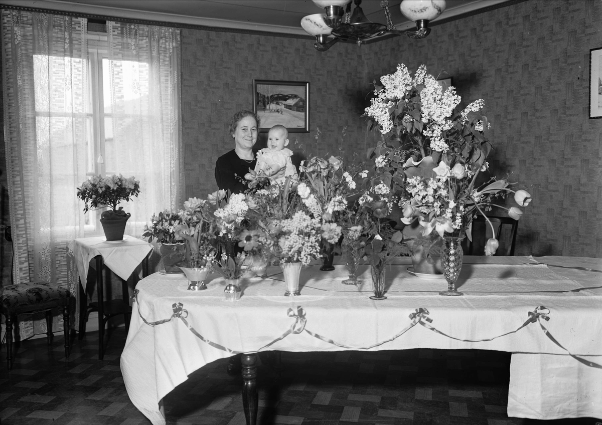 En kvinna som håller i ett barn som är iförd dopkläder står vid ett bord prytt med flera blombuketter, Uppsala