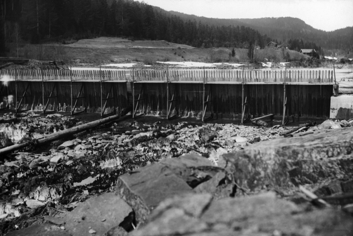 Såkalt «fermettedam» ved Tinnfoss i Tinnåa i daværende «Hiterdal» eller Heddal kommune, seinere i Notodden kommune.  Det er grunn til å anta at fotografiet er tatt tidlig på 1900-tallet.  Den første dammen av denne typen skal ha vært konstruert av en fransk ingeniør ved navn Poirée i 1831.  Poirées dam var utført med stålbukker som var hengslet til et fundament, som også var av stål.  På den øvre delen av bukkene var det anlegg for ei gangbru, som var sammensatt av elementer hadde en lengde som tilsvarte avstanden mellom bukkene, vanligvis om lag halvannen meter.  På motstrøms side av bukkene var det ei horisontal stålskinne, som «nålene» ble satt mot.  Dette var planker, gjerne i om lag 2" X 4" dimensjoner.  De ble stukket mot strømmen, slik at kreftene i vannet presset den nedre enden mot en terskel på damstedet.  Når nålene var presset tett sammen, sivet bare tynne stråler av vann gjennom damkonstruksjonen.  Dette fotografiet viser en slik fermettedam i nærheten av Tinnfossen i Tinnåa.  Fotografiet er tilsynelatende tatt tidlig på våren, mens det var liten vannføring i elva og mens det ennå lå snøflekker på bakken.  Til høyre i bakgrunnen ser vi et lite gardsbruk på en bakkekam mot Tinnåa.  Ellers preges bakgrunnen av barskogkledde åser.