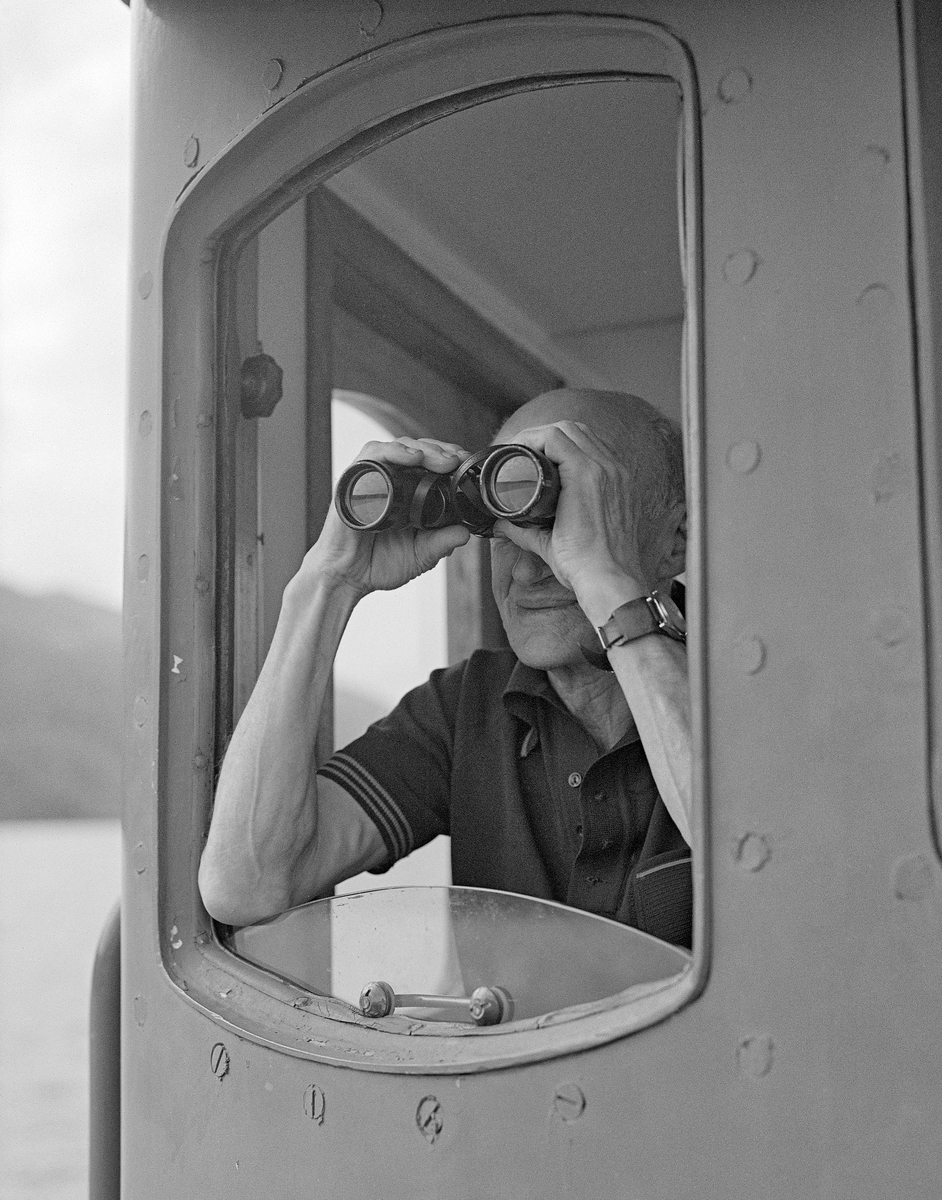 Rendølen Sigvart Svalstad (1915-2003), fotografert mens han speidet ut av et av vinduene i styrhuset på slepebåten «Storsjø» på innsjøen med samme navn i Ytre Rendalen i Hedmark.  Kikkerten var et nyttig hjelpemiddel, både når en skulle se at ringbommen bak båten var intakt, og når man, som her arbeidet med sluttrensk og skulle ha med seg alle restbeholdninger som lå og fløt langs strendene.  Sigvart Svalstad ble tatt inn i besetningen på slepebåten Storsjø i 1942, da under ledelse av sesongansatte sjøfolk fra Østfold.  Etter at dampmaskinen ble erstattet av en dieselmotor i 1957-58, var det lokale folk under ledelse av veteranen Halvor Fiskvik (1908-1989).  Etter at Fiskvik ble pensjonist fikk Svalstad gleden av å være skipper de siste tre sesongene det foregikk tømmerfløting på Storsjøen.  Båten hadde da en besetning på fire mann, skipperen inkludert.