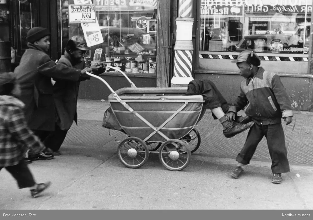 En grupp barn leker med en barnvagn i vilken en utslagen man ligger. Lenox Avenue, Harlem, New York, USA.