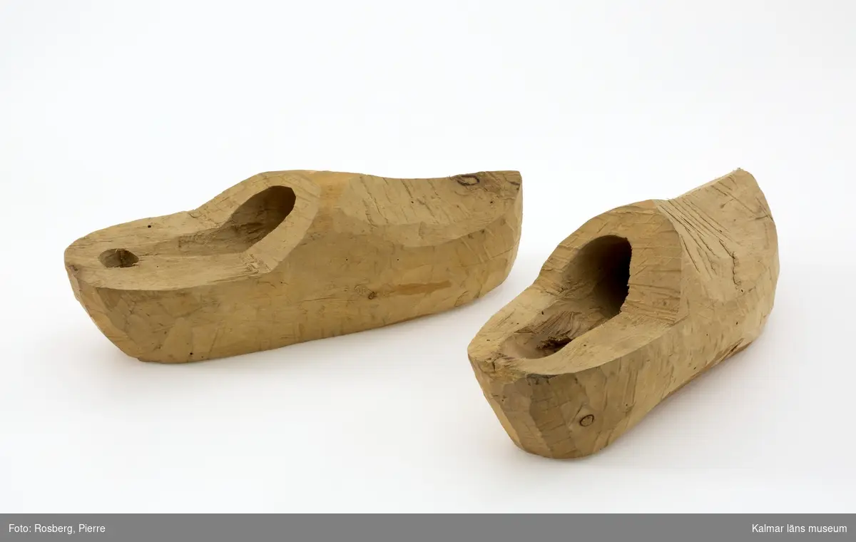 KLM 10032:1-2. Skor, träskor, halvfabrikat. Ett par. Tillverkade av träskomakaren för att visa på olika stadier i arbetet. Av trä. Utsidan är grovt tillhuggen med yxa. Klacken är ej utsågad. Insidan har en smalt, borrat hål. Sko :1 har ett mindre hål än sko :2. På sko :2 är dessutom hälpartiet grovt tillhuggen.