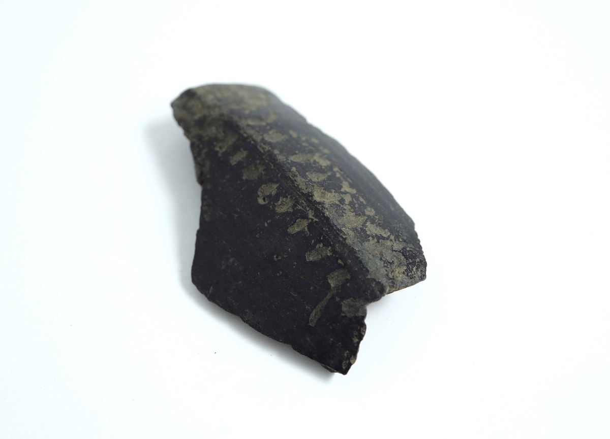 En bit av skuldran från ett gråbrunt, keramiskt gods med svart, blankglättad in- och utsida. Sannolikt del av någon form av kruka. Enkel dekor av lodräta linjer på var sida om skuldran. Tillverkad i Nederländerna under 1700- eller 1800-talet vid en tid då man ville efterlikna romersk keramik. Sammanhör med M 17000-2571 och 2572.