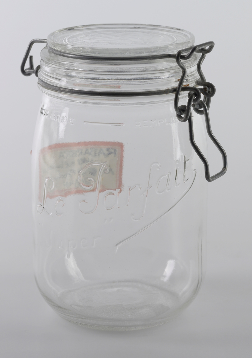 Et oppbevaringsglass laget av blankt glass med  glasslokk. Lokket er festet til glasset med ståltråd med en åpne-/lukkemekanisme. Det er tekst i relieff på undersiden av lokket, på siden av glasset og i bunnen, se "Påført tekst/merker". Det er også limt på en papiretikett med påskrift fra 1982, se "Påført tekst/merker". På undersiden er glasset ruglete. Glasset er av merket "Le Parfait Super".