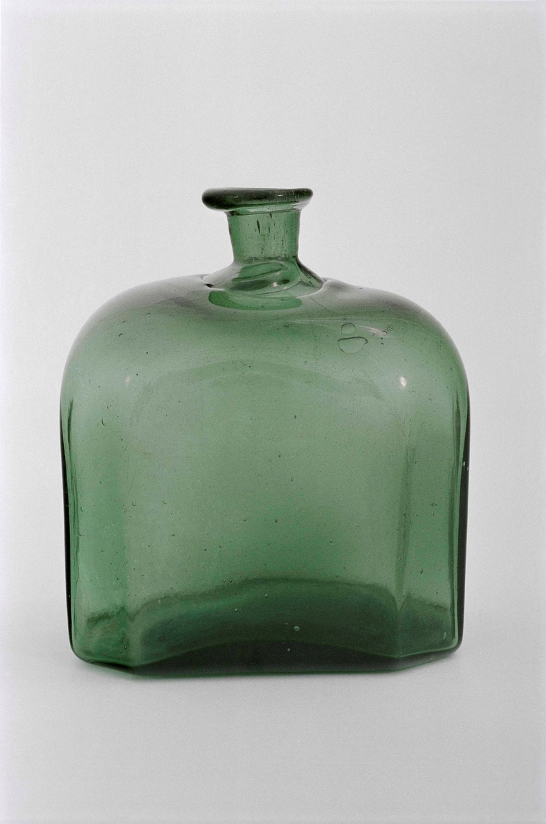Flaska av grönt glas, åttakantig, låg och bred med kort hals.
Halsen med kraftig fals.