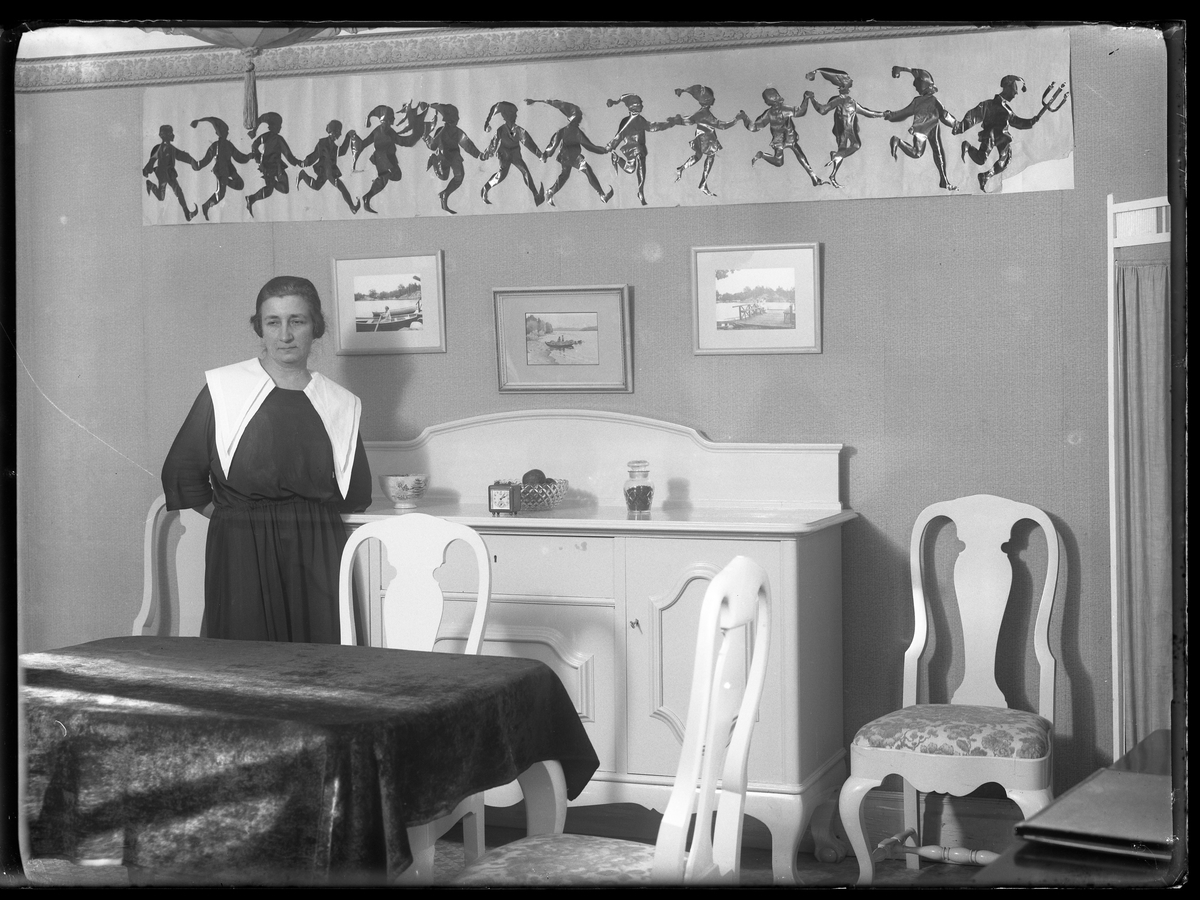 Anna Lenander står lutad mot en vit skänk i en matsal. På väggen hänger en pappersbonad med tomtenissar i dans.