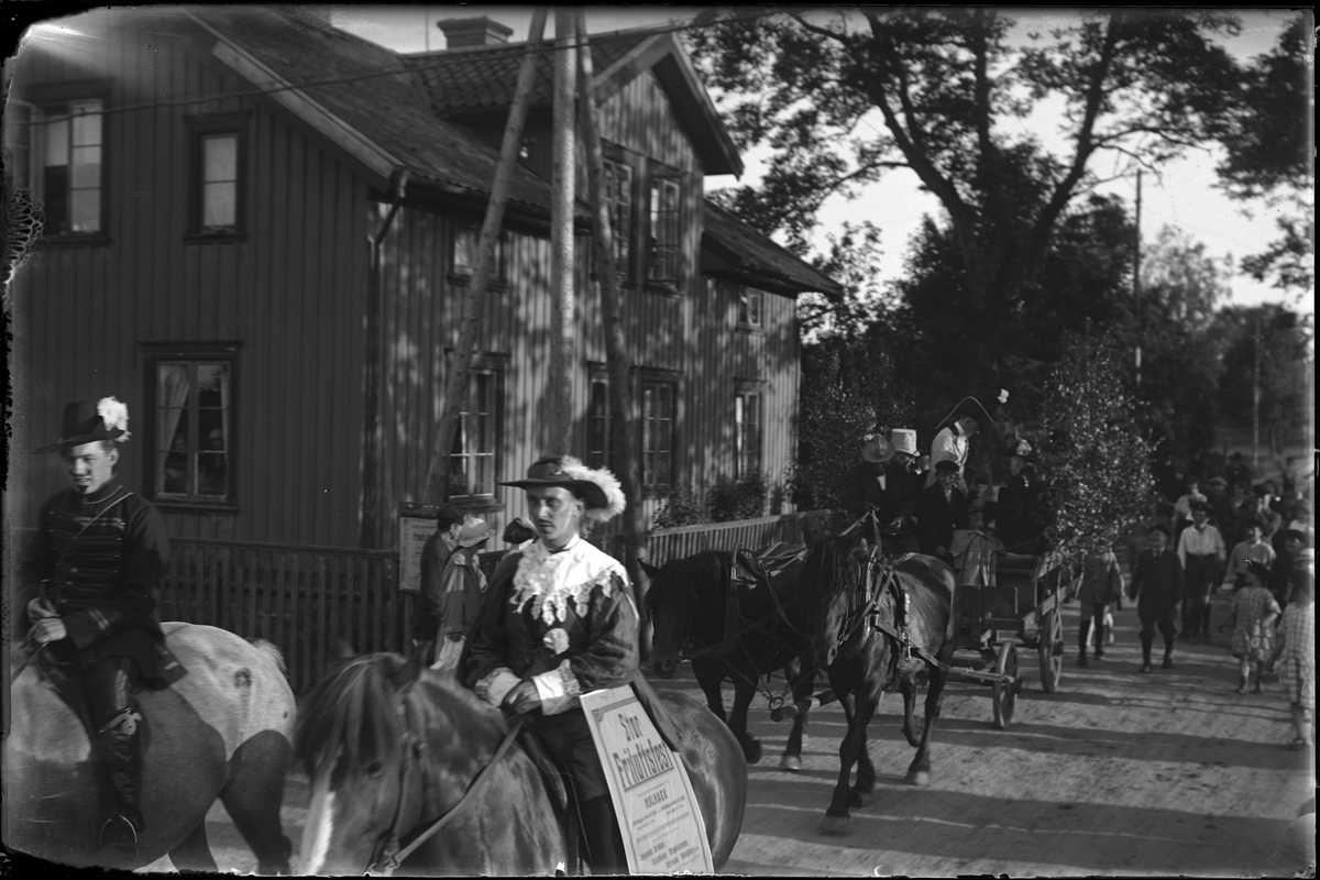 Två utklädda män kommer ridande, efter dem kommer två hästar med vagn samt ett antal promenerande människor. I fotografens egna anteckningar står det "Jazzkapellet A.I.F.s fest"
