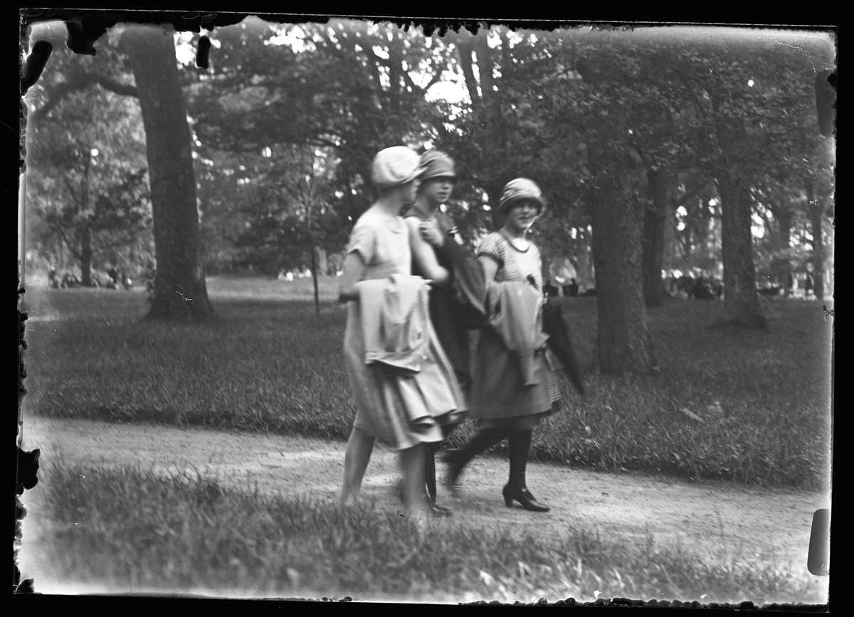 Tre sommarklädda kvinnor promenerar i Nolhaga park. I bakgrunden sitter ett antal personer i gräset mellan träden.