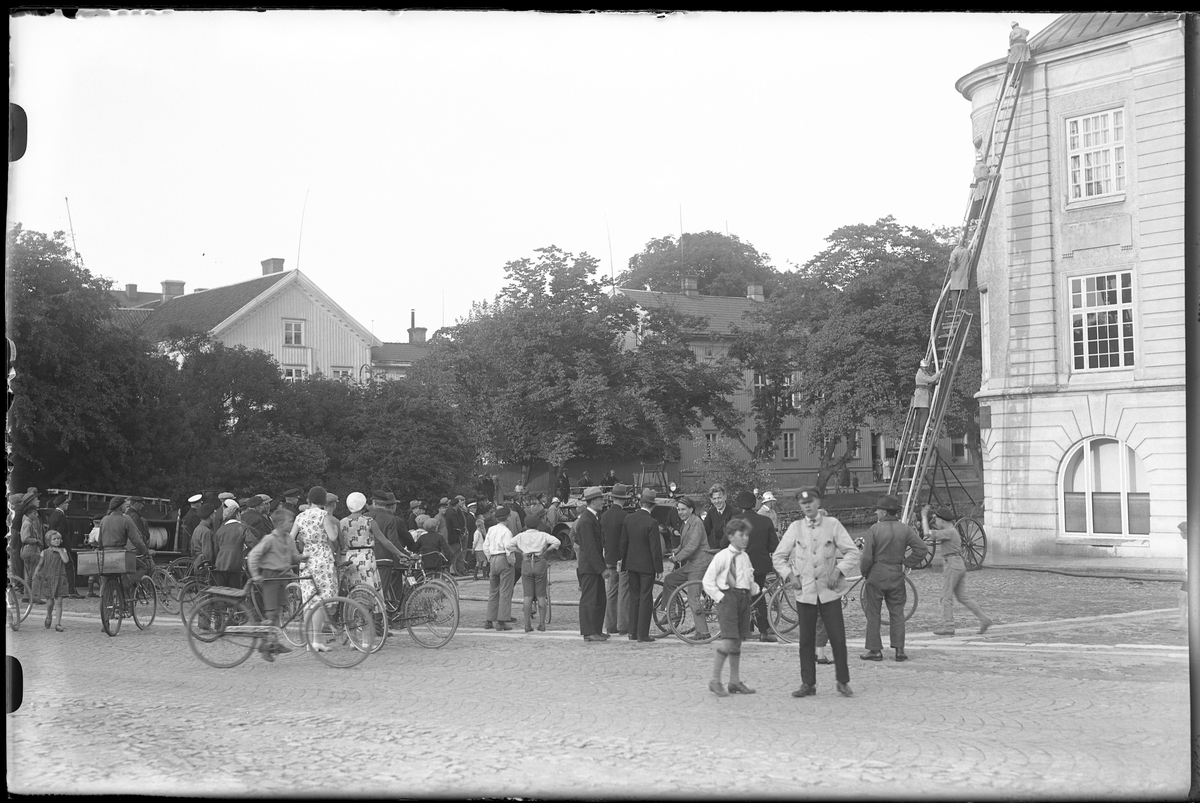 Fyra brandmän står på en stege. Stegen står lutad mot gamla Sparbankshuset på Lilla torget och nere på torget står ett stort antal åskådare och tittar.