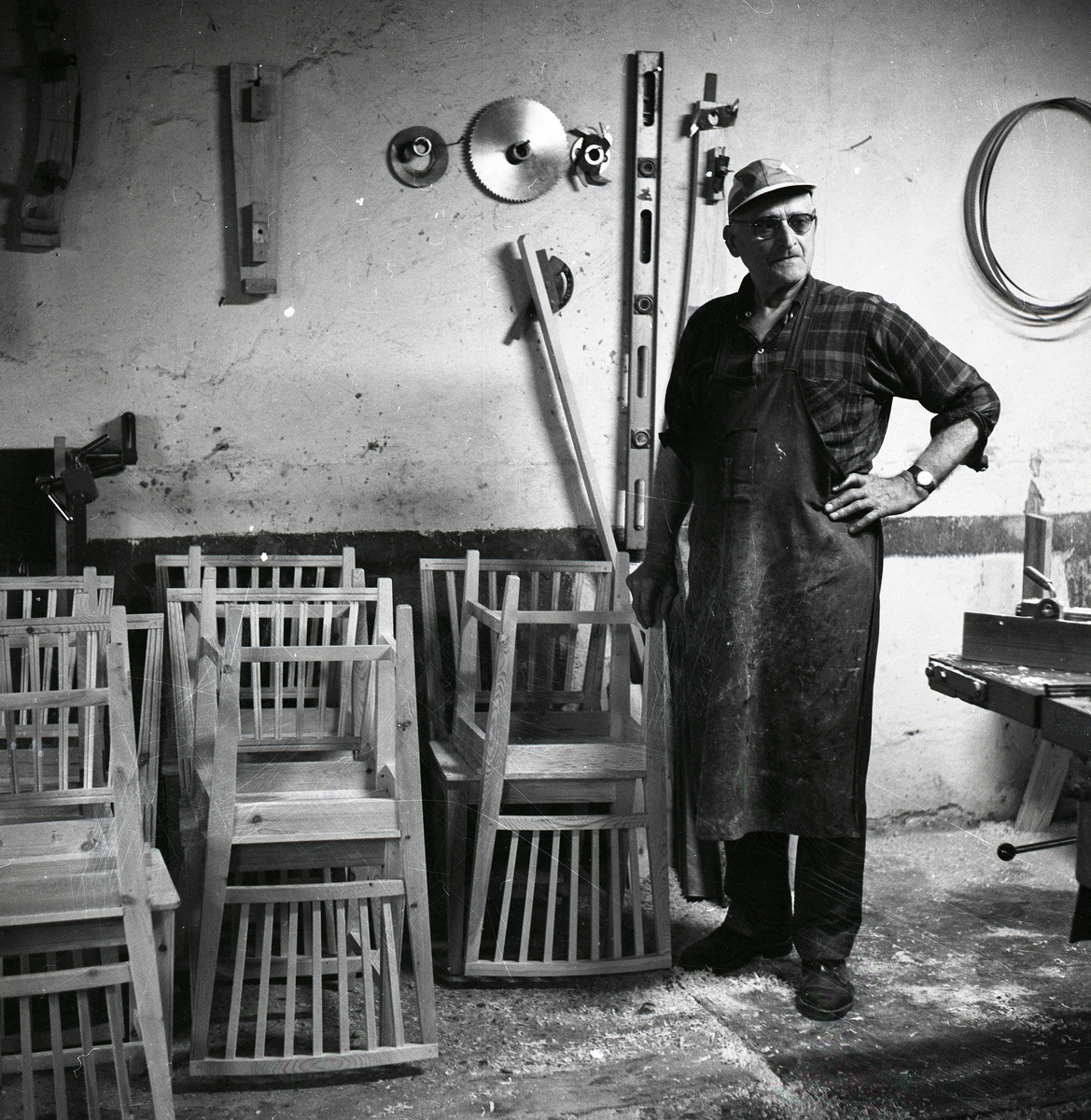 En man i förkläde och keps står bredvid några stolar i en verkstad, 1968.