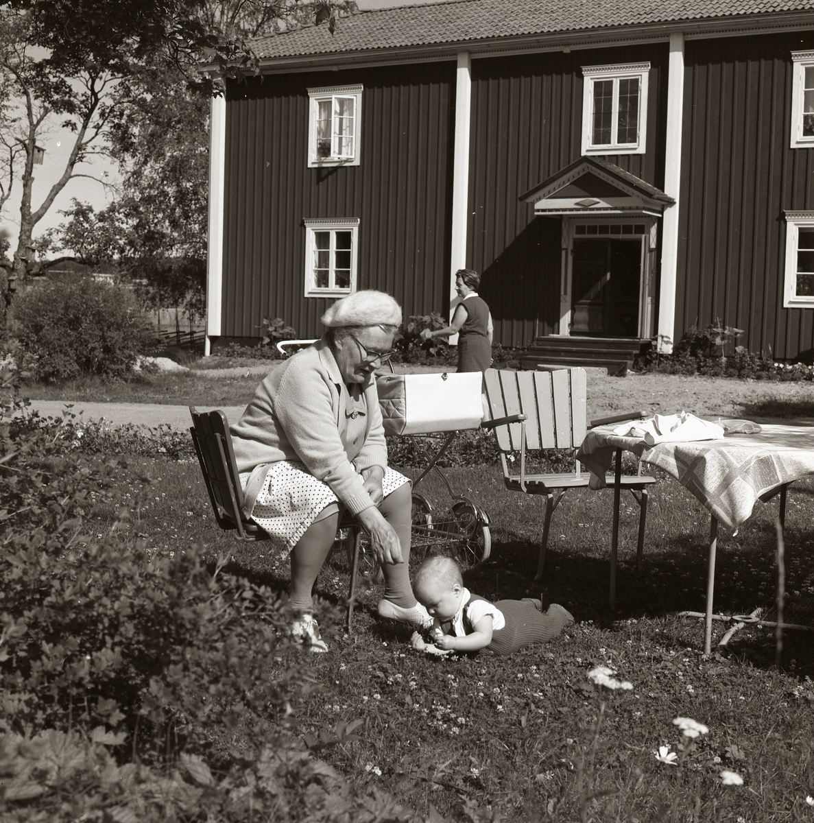 En kvinna sitter på en trädgårdsstol och passar ett litet barn som ligger på marken framför henne. I bakgrunden syns bostadshuset på gården Sunnanåker, 1970.