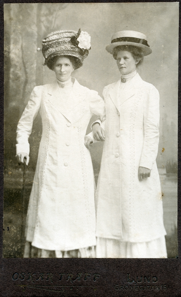 Ateljéporträtt av Mariana Träff och Emilia. De har båda stora hattar och är iklädda knälånga ljusa kappor samt långa kjolar. Mariana håller en käpp i ena handen. Kvinnorna står armkrok.